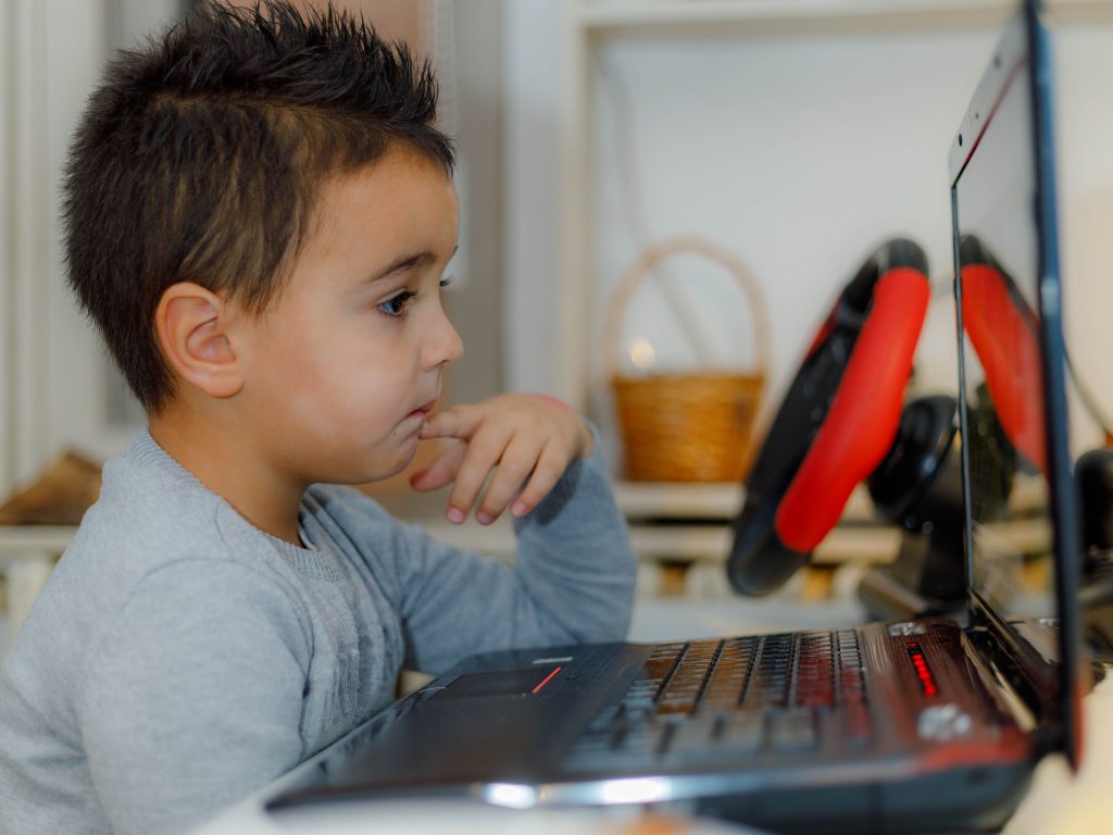 A young boy focuses intently on a laptop, surrounded by gaming accessories indoors.