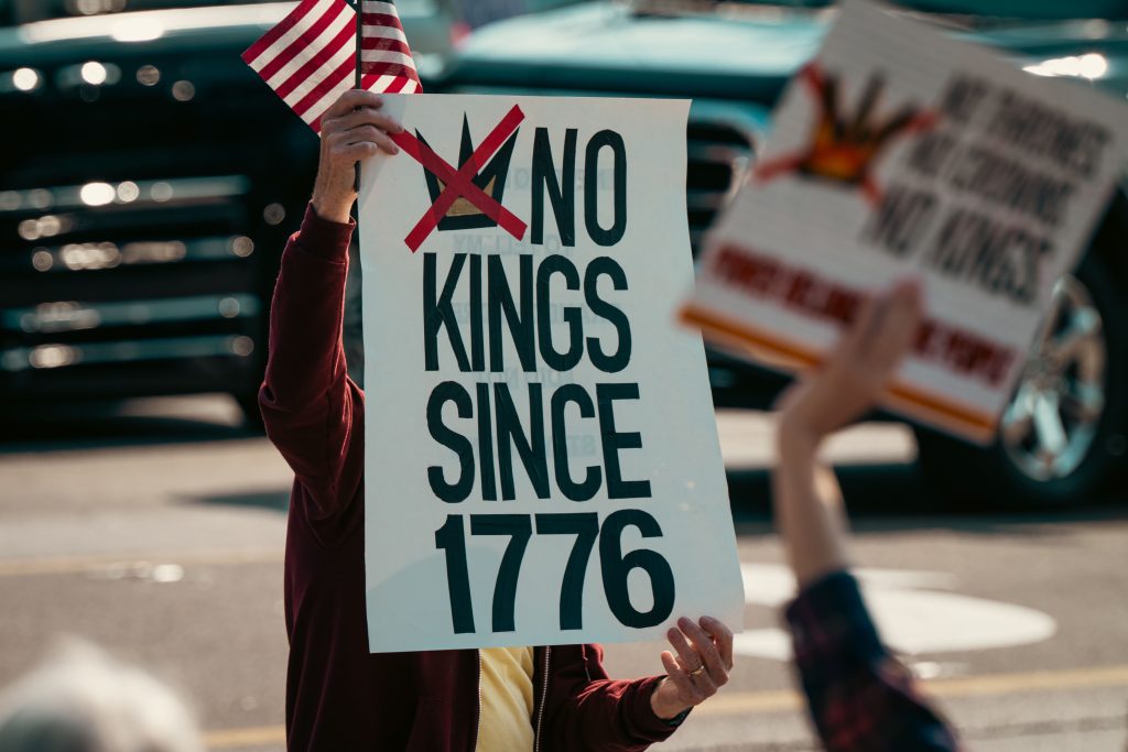 Protest sign displaying 'No Kings Since 1776' held alongside a small US flag, emphasizing democratic sentiments.