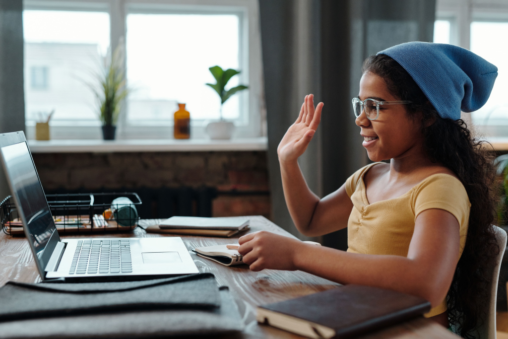 A young girl waves at her laptop during a video call in a cozy home office setting.