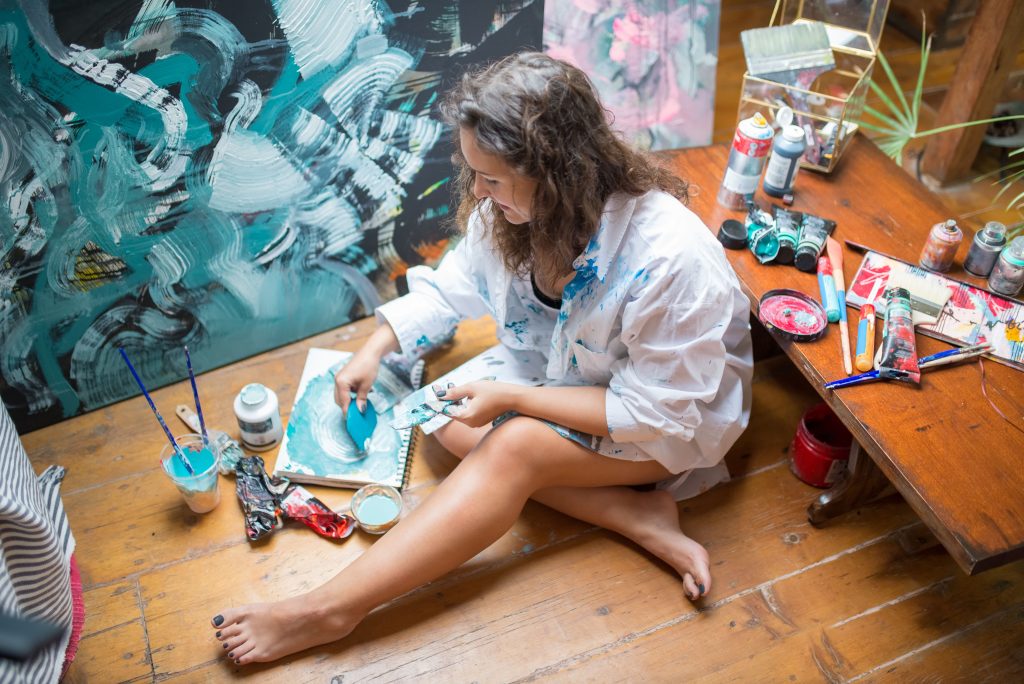 A woman artist painting abstract blue artwork indoors, surrounded by art supplies.