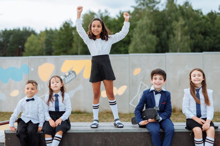 A diverse group of cheerful children wearing school uniforms, smiling and celebrating together in an outdoor setting.